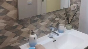 A bathroom with a white sink, a mirror above it, and a patterned wall. There are two towels on the sink, a soap dispenser, and a bottle of hand soap. - Private new flat (Bari)