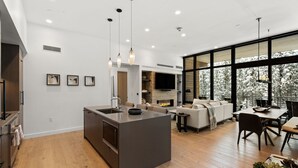 A modern kitchen with a central island, a dining area with a table and chairs, a living room with a sofa and fireplace, and large windows with a view of trees. - Luxury Moonlight Basin 2 Bedroom Condo (Big Sky)