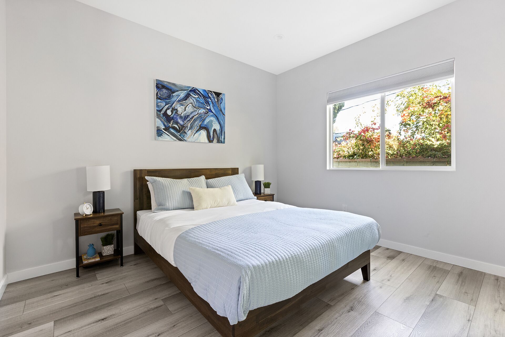Una camera da letto con un letto, due comodini, un tavolino, una finestra vista vegetazione e un quadro appeso al muro.