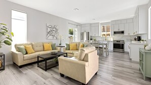 Ein modernes Wohnzimmer mit einem beigefarbenen Sofa, einem Couchtisch und einem Essbereich, wobei im Hintergrund eine Küche zu sehen ist.