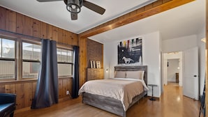 Un dormitorio con suelo de madera, una cama con cabecera gris, un ventilador de techo y un ventanal con cortinas.