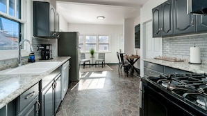 Cocina moderna con encimeras de granito, estufa a gas y muebles blancos. Cuenta con una zona de comedor con mesa y sillas, y un ventanal que proporciona luz natural.