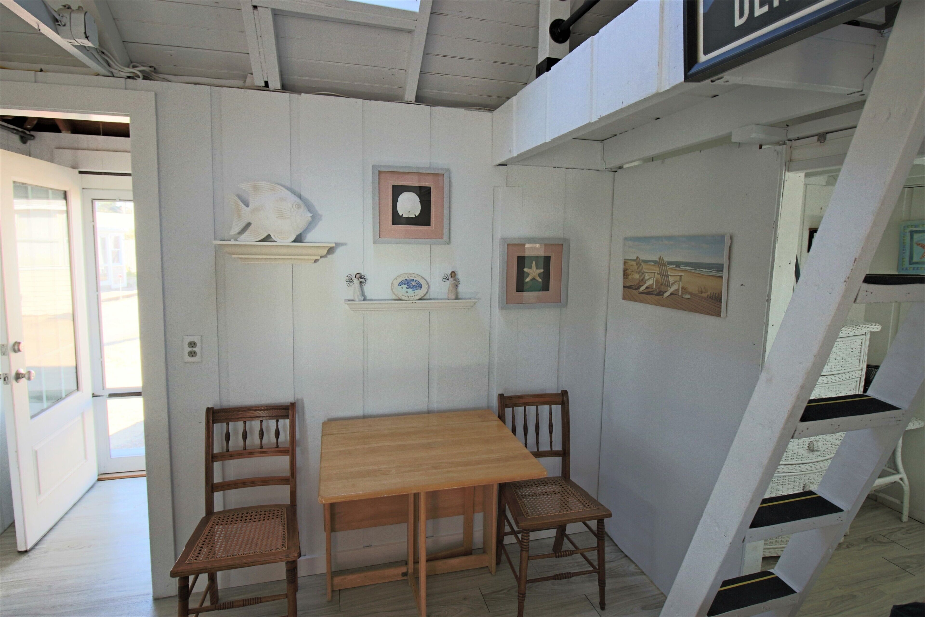 Une table à manger en bois avec deux chaises, un mur blanc orné de tableaux encadrés et un escalier.