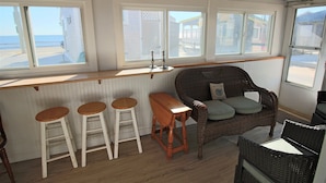 A room with a wooden ceiling, a wicker sofa with cushions, a wooden bar stool, and a view of the beach through large windows.
