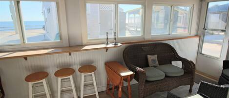 Una habitación con techo de madera, un sofá de mimbre con cojines, un taburete de bar de madera y vistas a la playa a través de amplias ventanas.