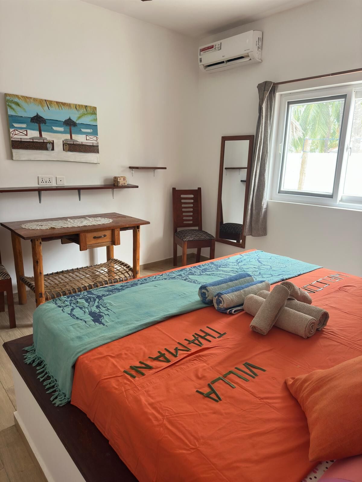 A bedroom with a bed, a desk, a chair, a painting, and an air conditioning unit.