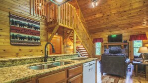 A wooden cabin interior with a kitchen, dining area, and a staircase leading to a loft.