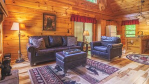 A cozy log cabin interior with a leather sofa, ottoman, and matching armchair, a dining area with a wooden table, and a kitchen with a refrigerator.