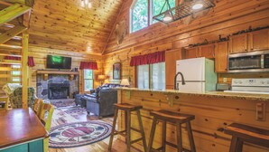 A wooden cabin interior with a stone fireplace, wooden dining table, bar stools, and a kitchen area with a microwave and refrigerator.