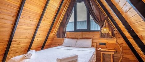 A cozy attic bedroom with a wooden sloped ceiling, a bed with white linens, a small wooden nightstand, and a window with curtains.