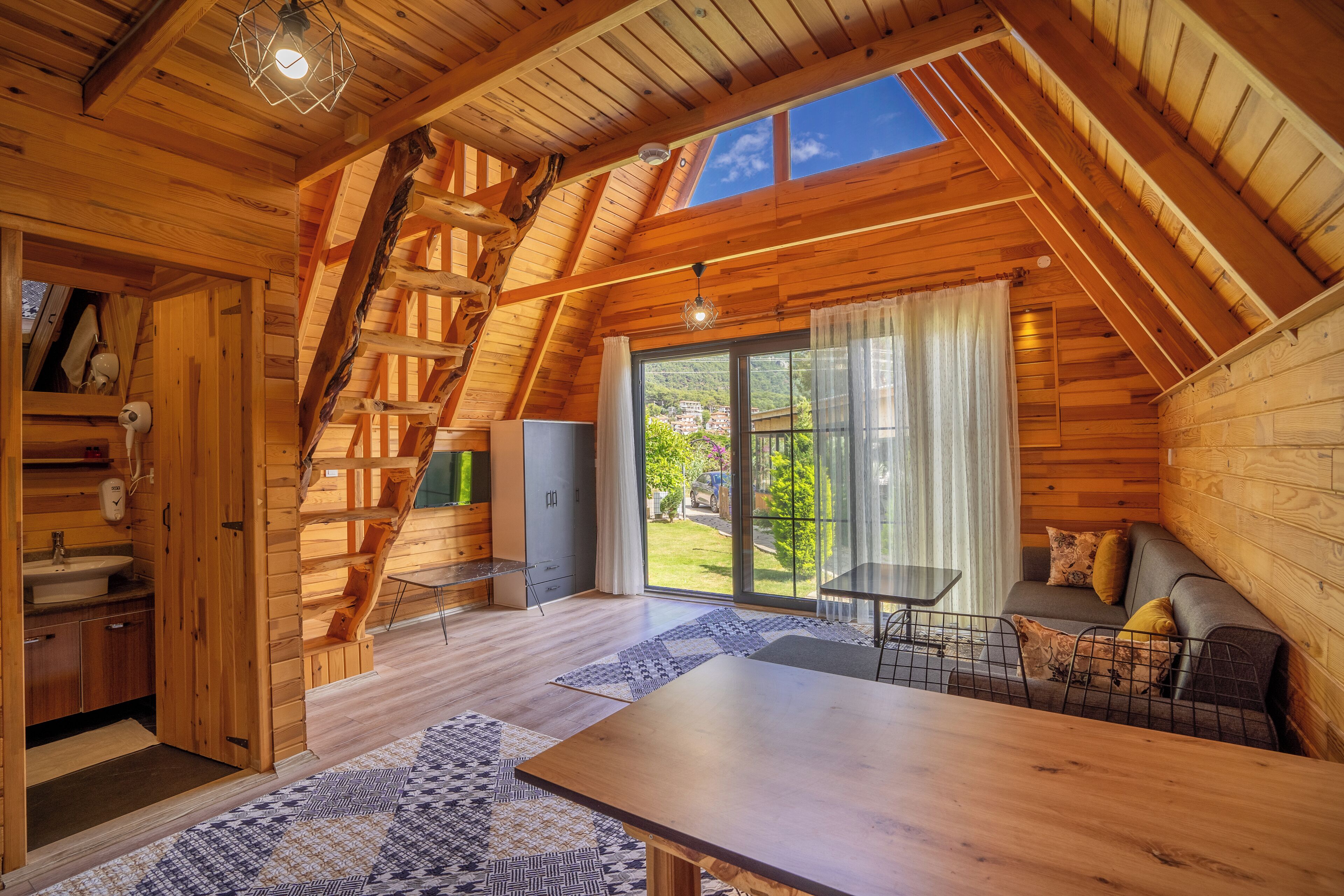 A wooden cabin interior with a dining area, a sofa, a staircase, and a large window offering a view of greenery.