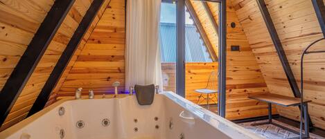 Una cabaña de madera con techo inclinado, una bañera grande y un amplio ventanal corredera que da a un área exterior.
