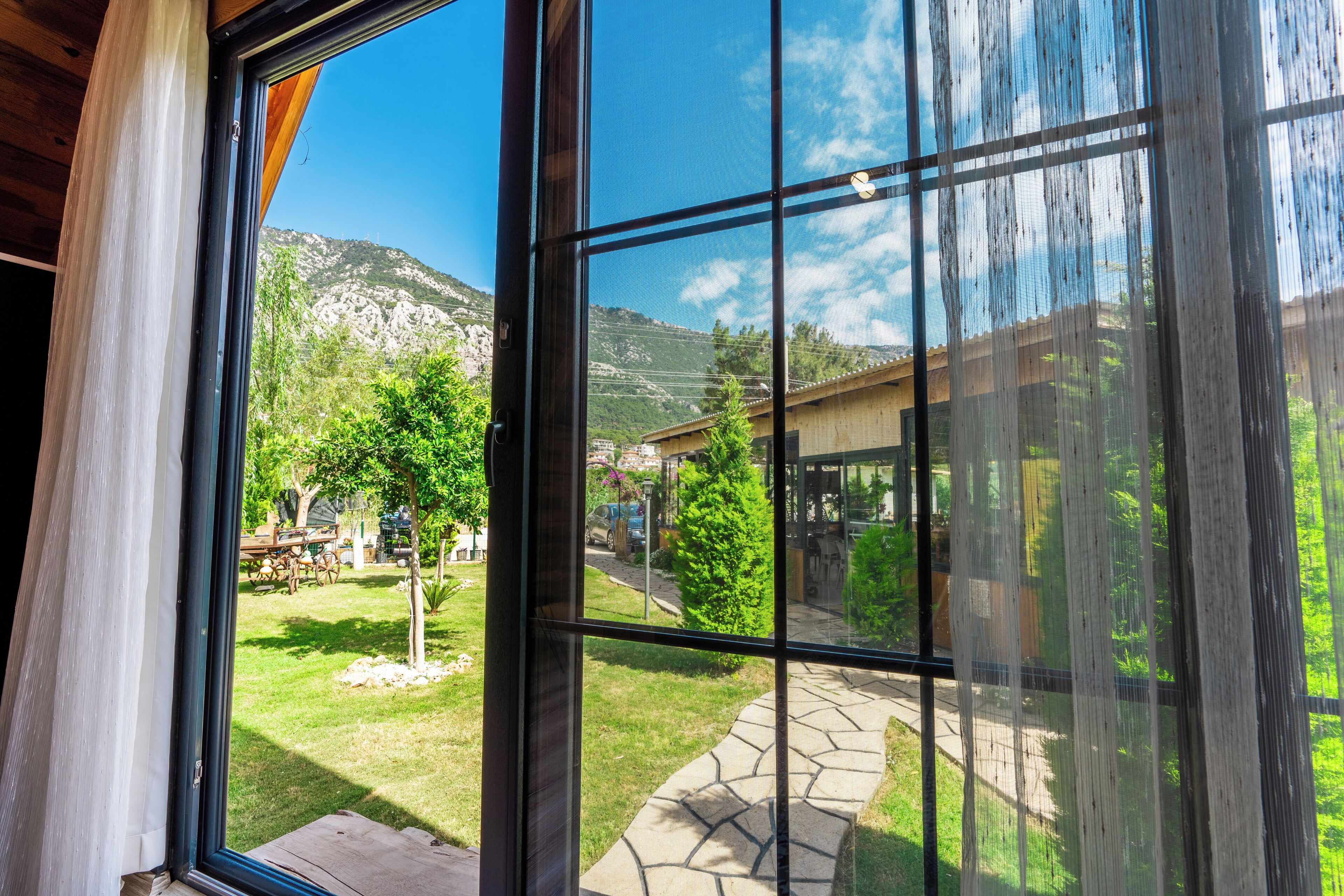 Blick durch ein Fenster auf einen gepflasterten Weg, der zu einem Gebäude führt, umgeben von Grünpflanzen und Bergen.