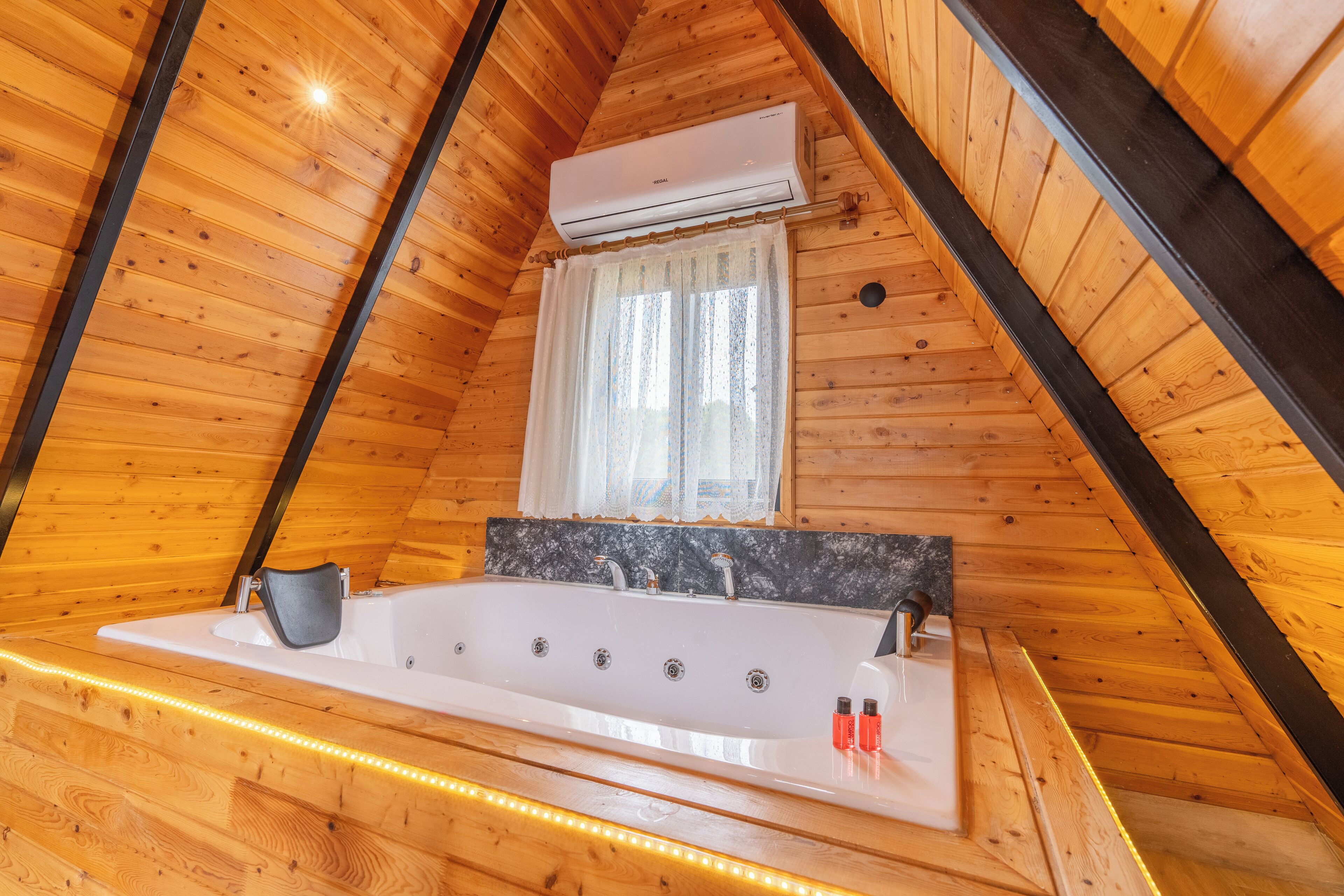 A wooden cabin bathroom with a bathtub, a window with curtains, and an air conditioning unit.