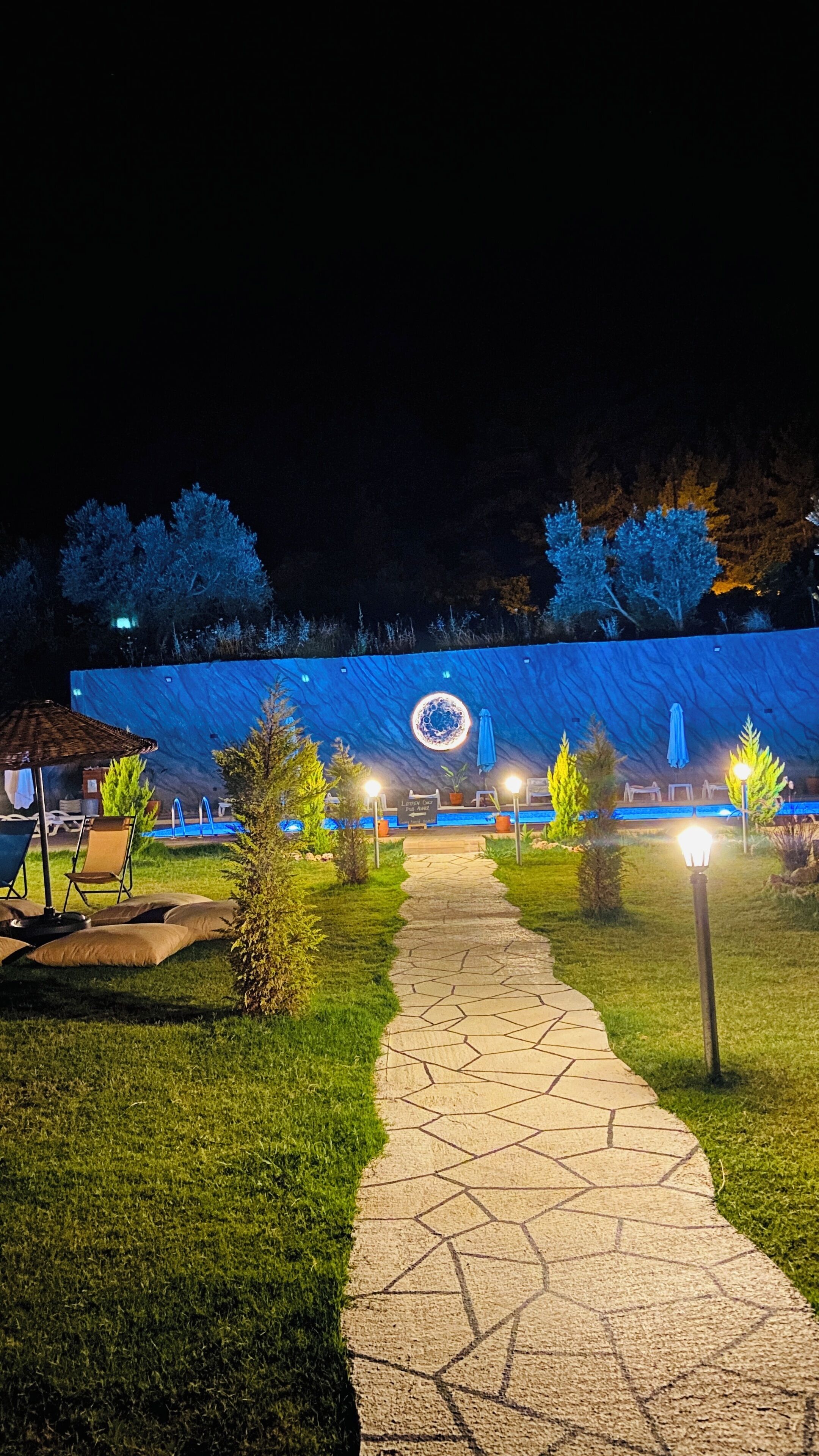 A pathway leading to a building with a blue facade and a circular emblem. There are outdoor seating areas and potted plants along the path.