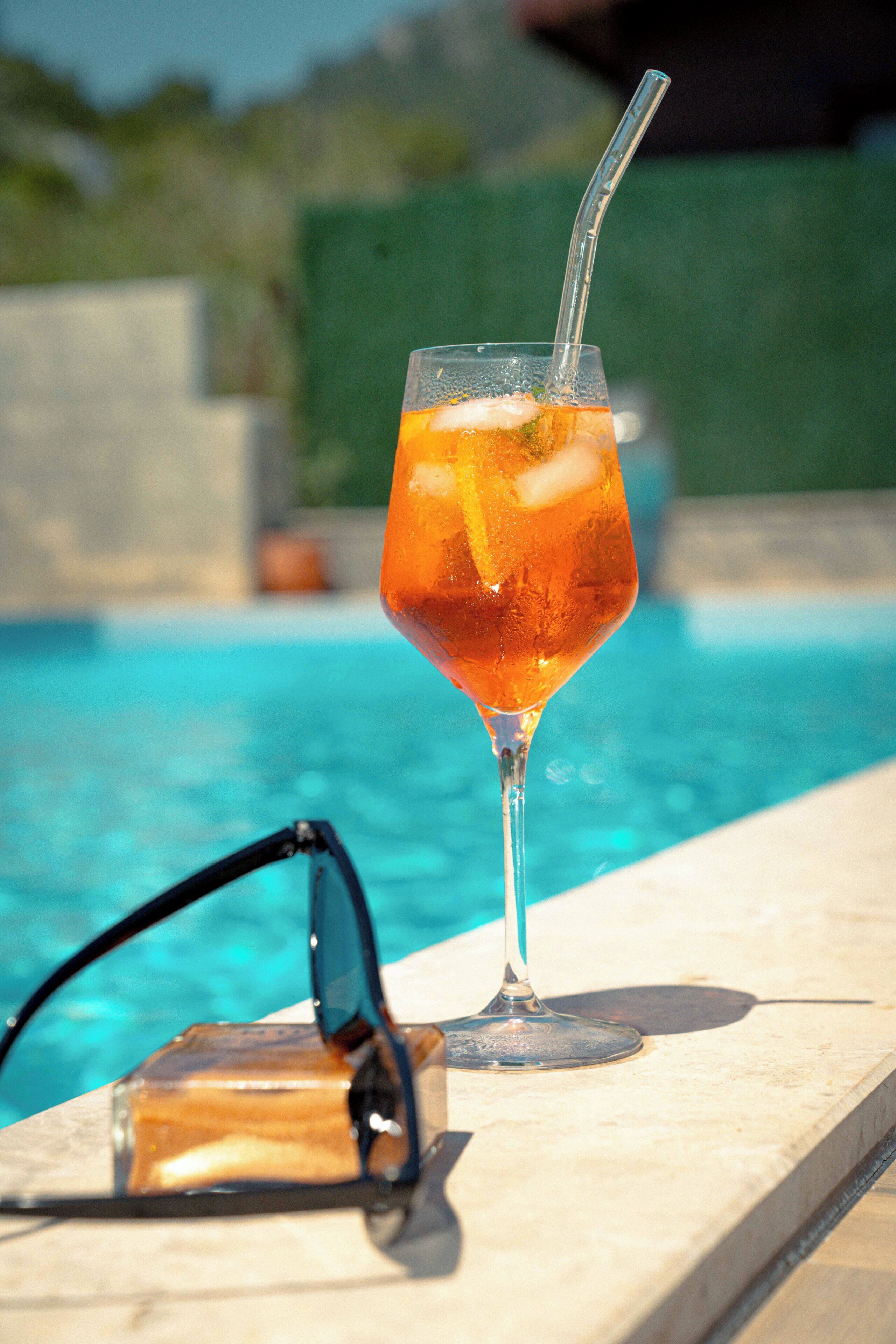 A glass with a straw and ice cubes next to a pair of sunglasses by a pool.
