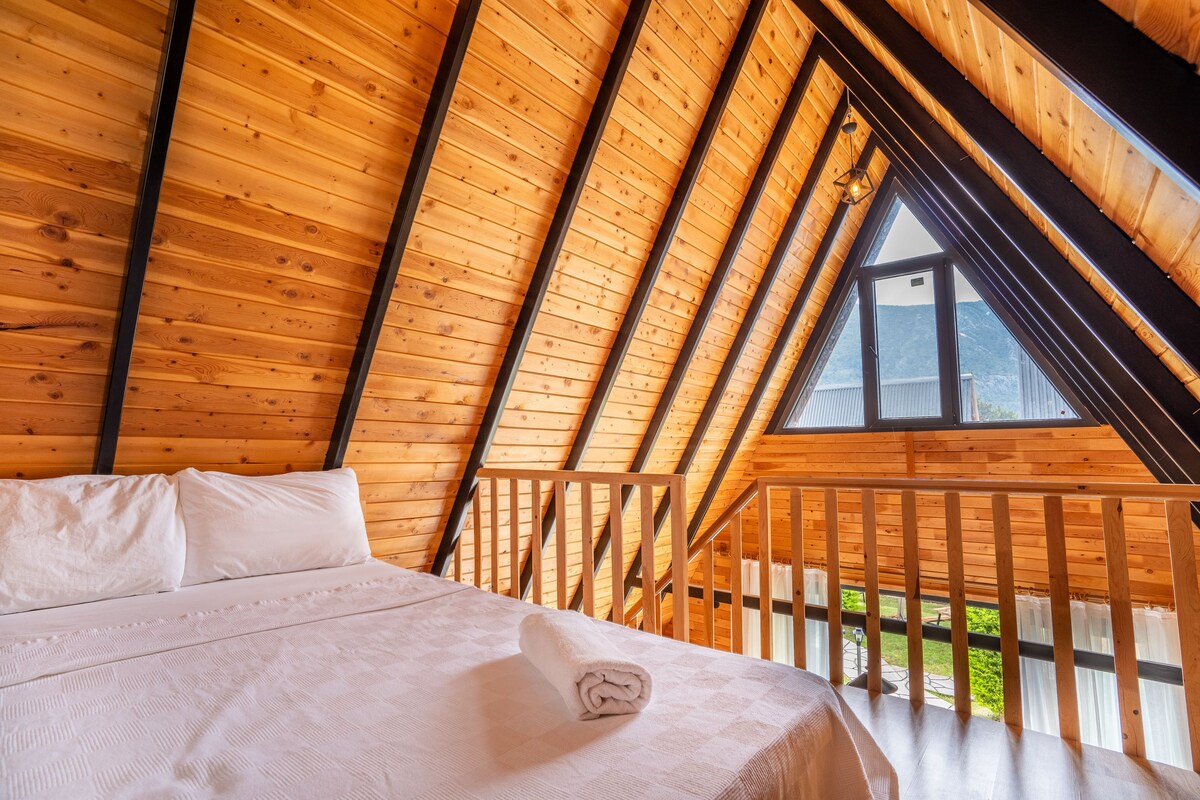 Ein Zimmer mit schrägverkleidetem Holzbalken, einem Bett mit weißer Bettwäsche und einem Blick durch das Fenster hinaus.