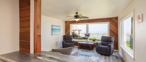 A spacious room with a wooden ceiling, a ceiling fan, a large window with a view of the ocean, and two leather chairs.