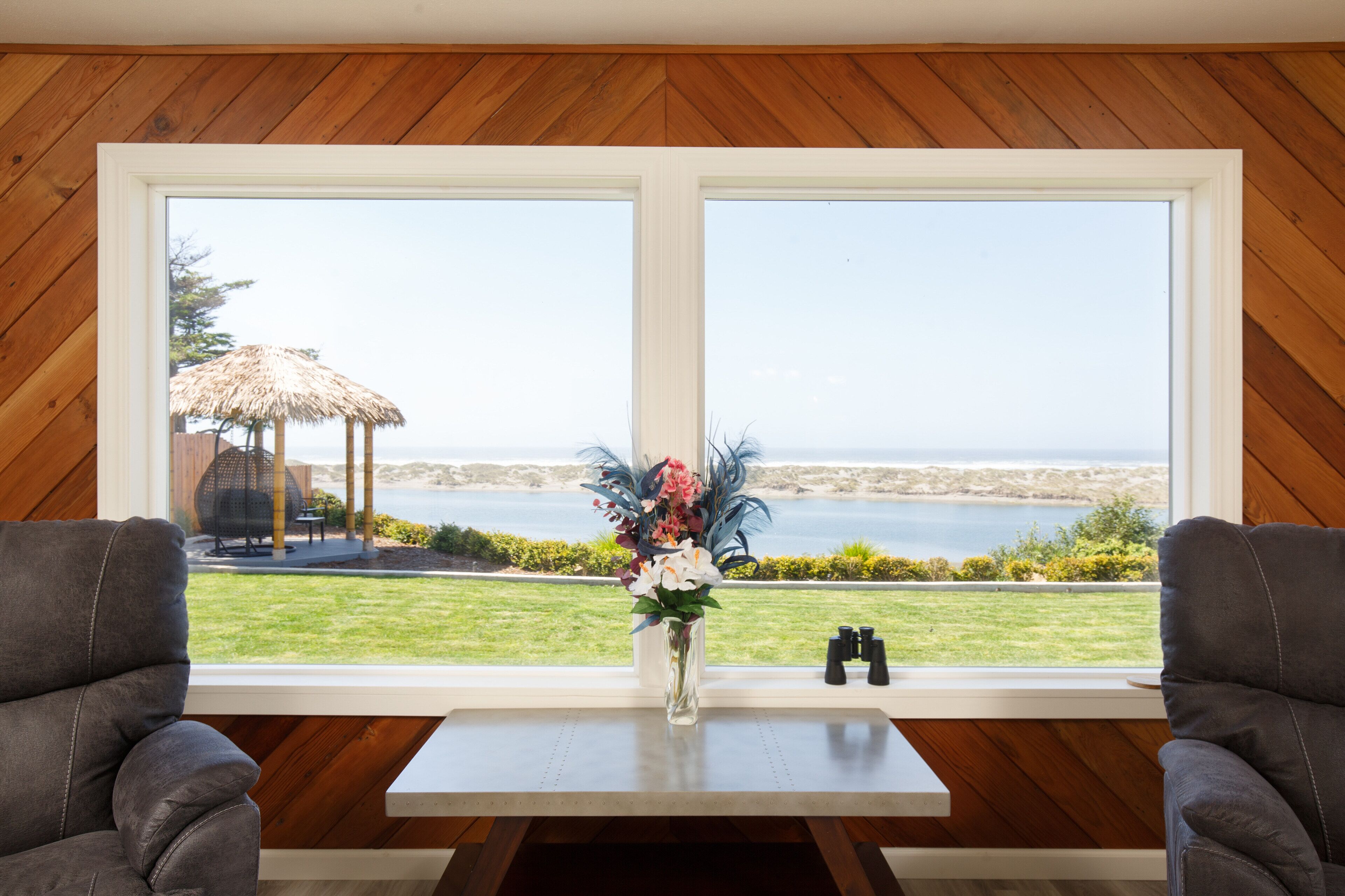 Une pièce dotée d’une grande fenêtre donnant sur une plage et la mer, d’un mur en bois, de deux fauteuils en cuir et d’une table sur laquelle est posé un vase de fleurs.