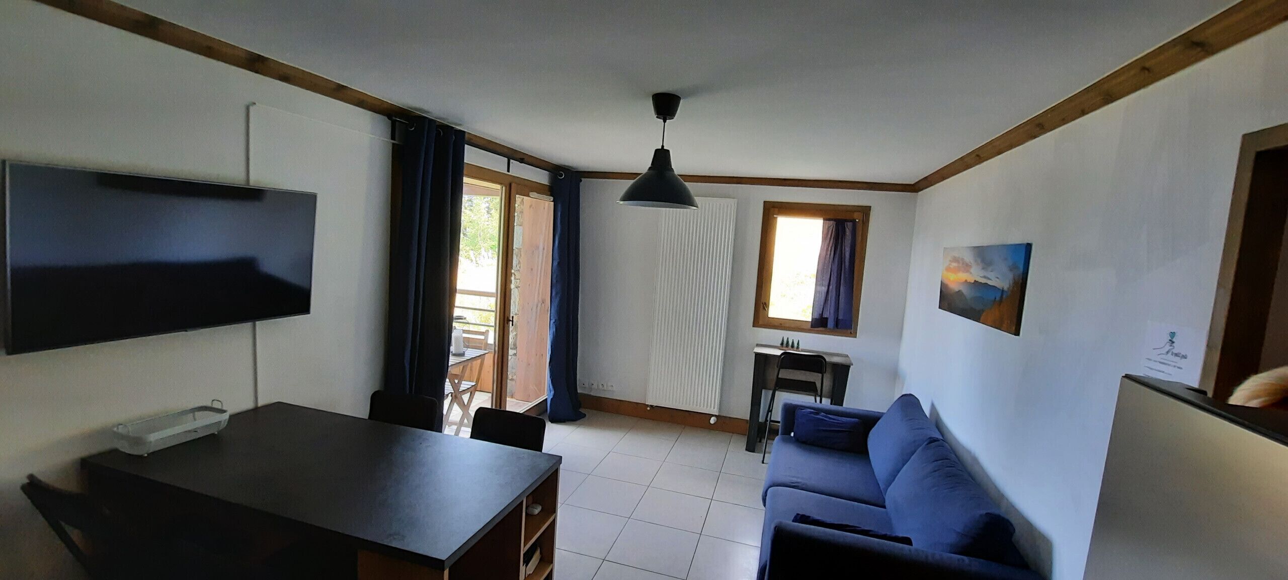 Ein Zimmer mit Fernseher, Tisch, Sofa, Schreibtisch mit Stuhl und Fenster mit Vorhängen.