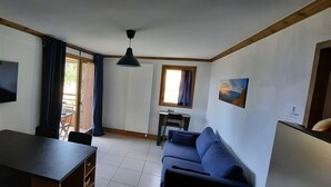 A room with a TV, a table, a sofa, a desk with a chair, and a window with curtains.