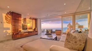 A modern living room with a large window overlooking the sea, a wooden accent wall, and a cozy seating area. - Beautiful New 3BR Condo at Indah (Puerto Vallarta)