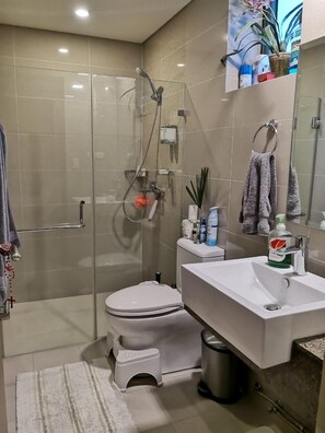 A modern bathroom with a glass-enclosed shower, a white toilet, a sink with a mirror, and a small trash bin. - Beautiful Corner Studio Near Resorts World Manila Across NAIA Terminal 3 (Pasay)