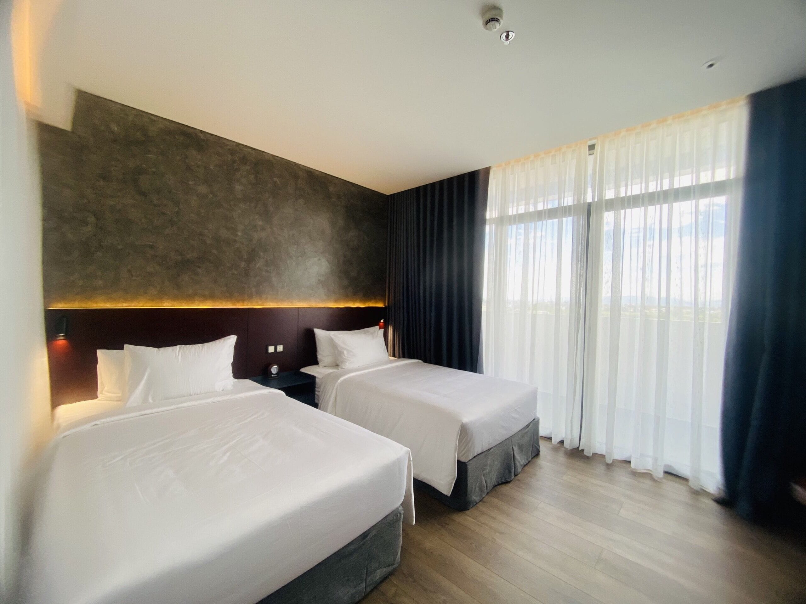 A hotel room with two beds, a large window with sheer curtains, and a modern headboard.