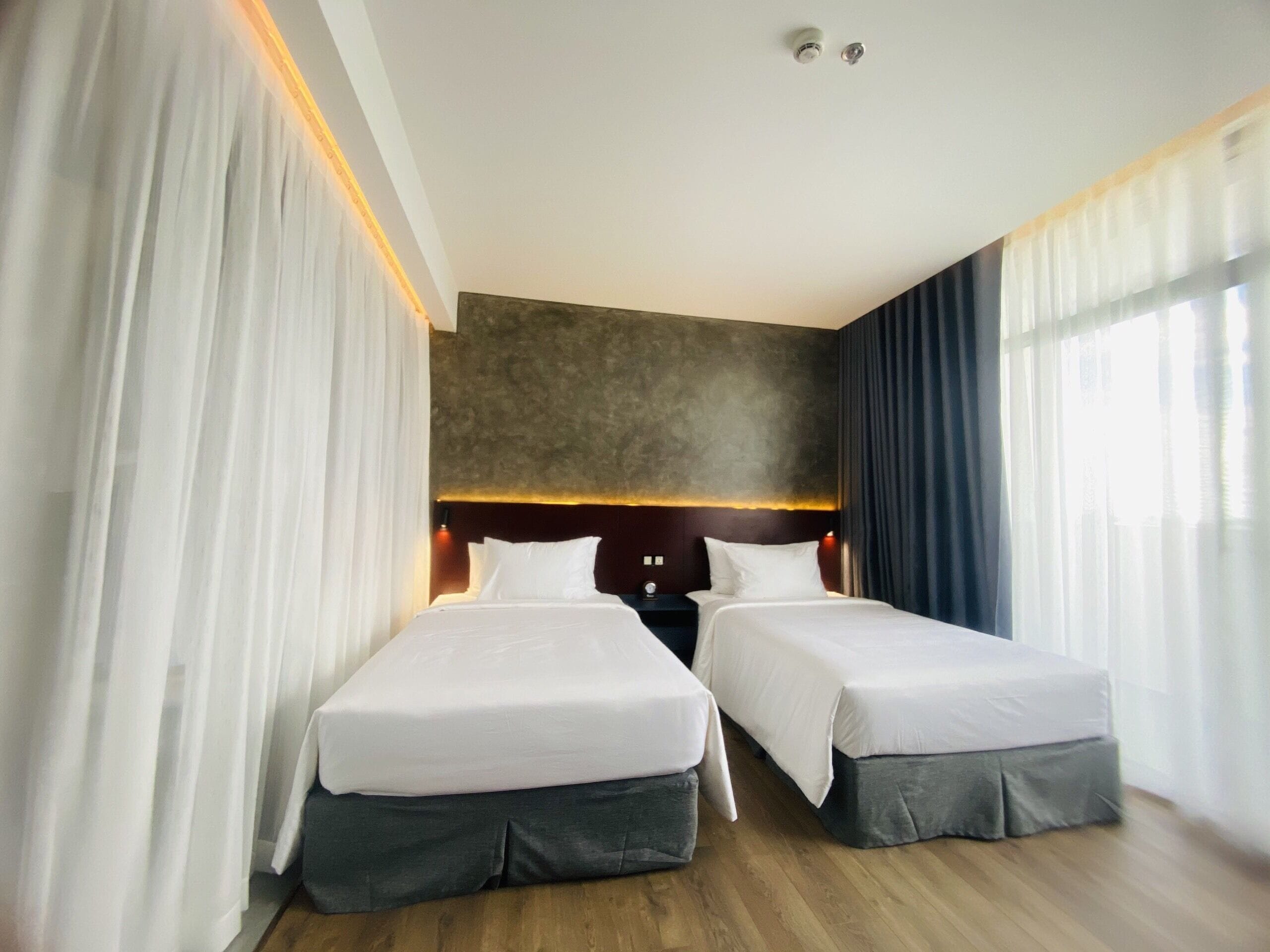 A hotel room with two beds, a wooden floor, a dark headboard, and a large window with curtains.
