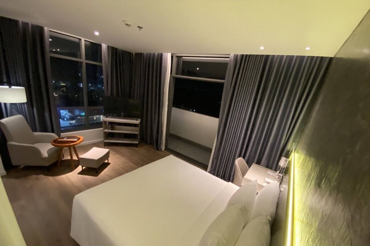 A hotel room with a large bed, a sitting area with a chair and a small table, and a view of the city at night through the windows.