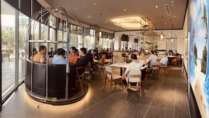 A modern restaurant with round tables and booths, large windows, and a central decorative light fixture. - NEWCC HOTEL & SERVICED APARTMENT (Truong Quang Trong)