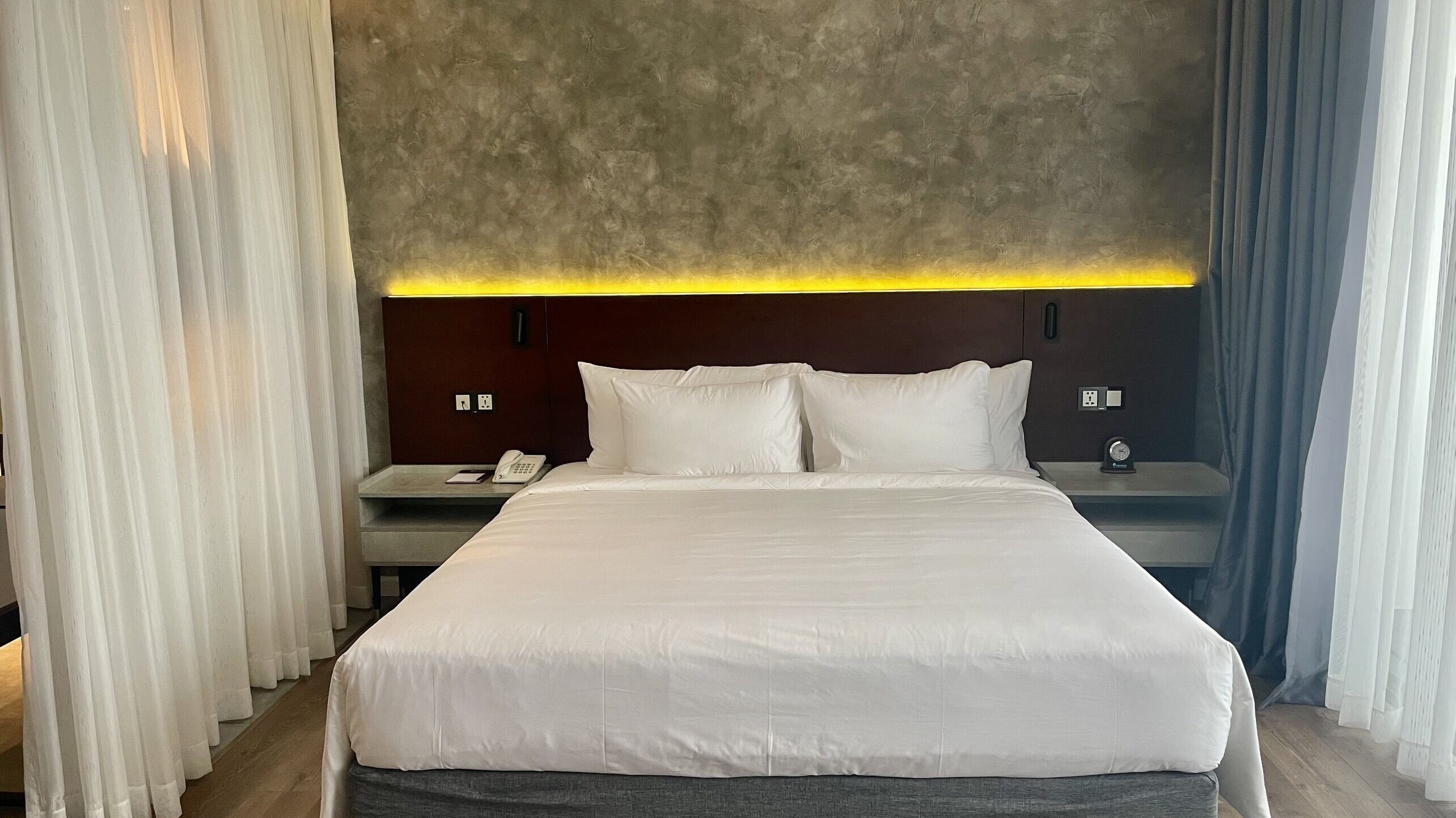 A modern hotel room with a large bed, a bedside table with a phone, and a nightstand with a clock.
