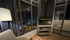 A modern hotel room with a large window offering a city view, a comfortable sofa, a wooden side table with a book, and a TV stand with a flat-screen TV. - NEWCC HOTEL & SERVICED APARTMENT (Truong Quang Trong)
