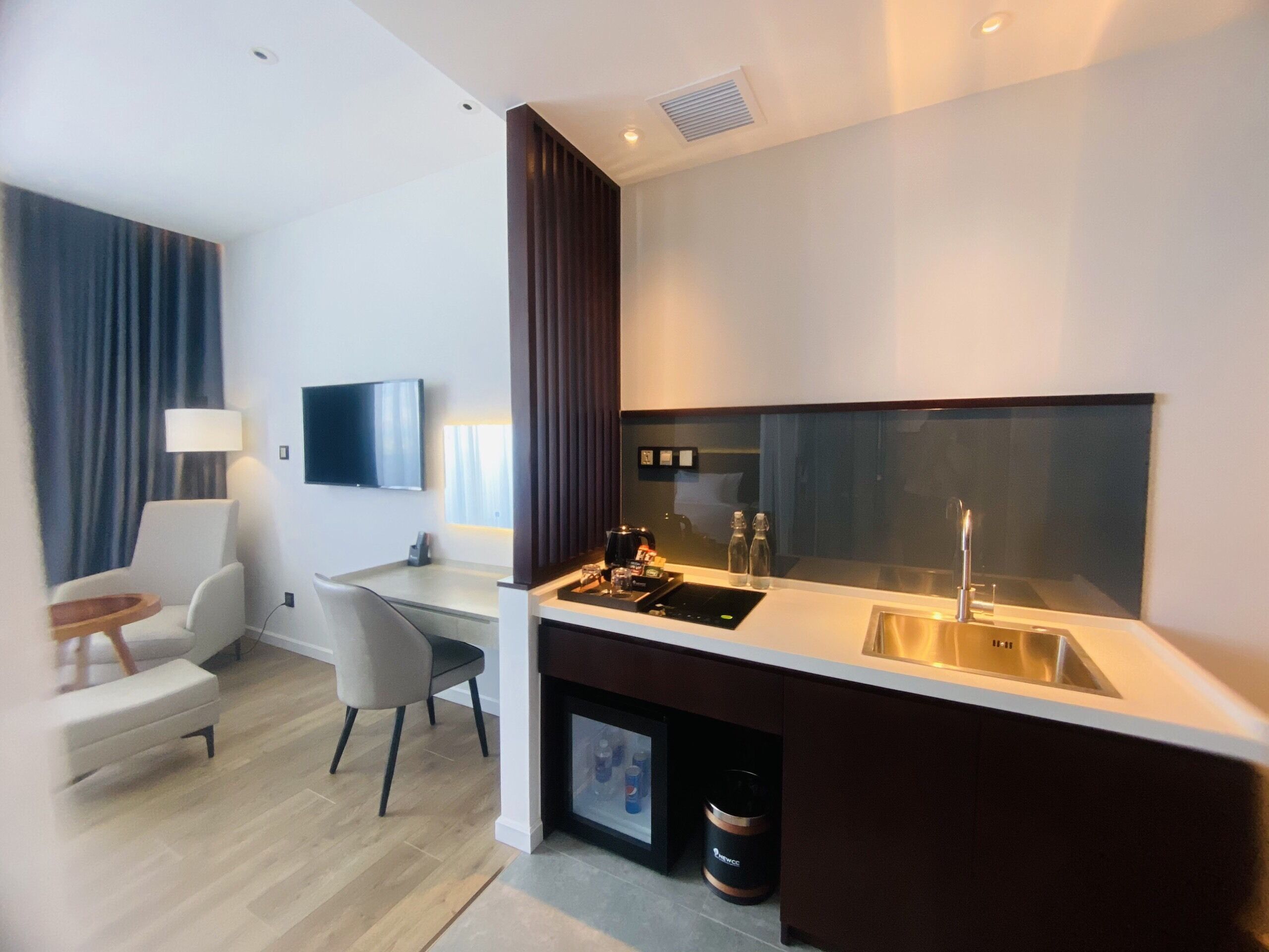 Quarto de hotel moderno com cozinha compacta, televisão de tela plana, área de estar com uma cadeira e mesa pequena, e uma janela grande com cortinas.