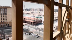 A view through a window with a geometric frame, featuring a parking lot with several cars, a street, and a large building with a distinctive architectural style in the background. - Sky View Hotel Madinah (Madinah)