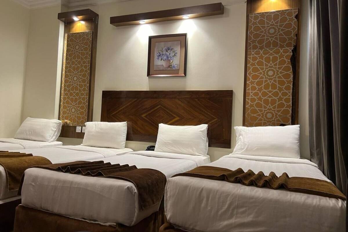 A hotel room with three beds, each with white linens and brown headboards. There is a framed picture on the wall above the beds, and decorative panels on the walls.