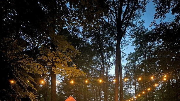 Une tente avec un feu devant, entourée d’arbres et illuminée de guirlandes lumineuses.