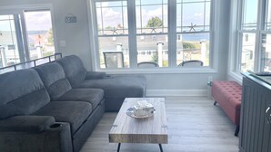 Ein Wohnzimmer mit grauem Sofa, einem Holztisch und einem pinken Pouf. Große Fenster bieten Blick auf das Meer und Gebäude.