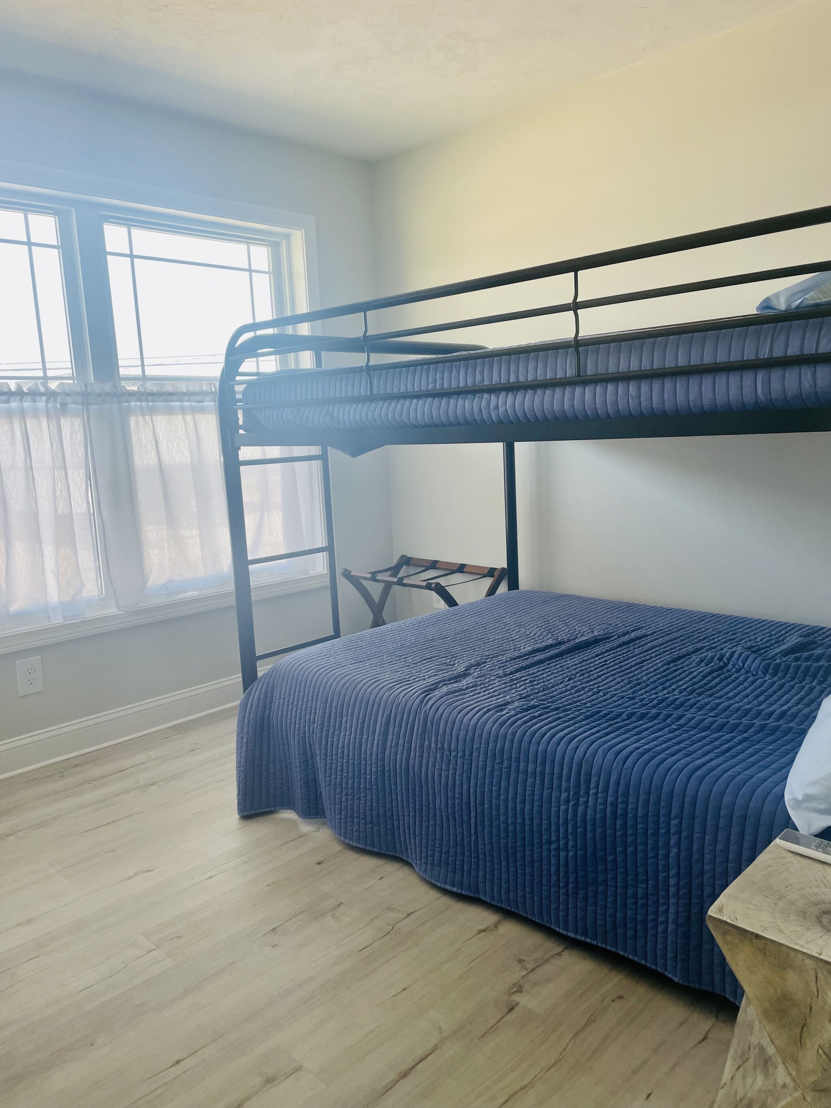 A bunk bed with a blue quilt, a small wooden table, and a window with curtains.