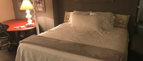 A neatly made bed with a white comforter and pillows, a red table with a lamp, and a framed picture on the wall.