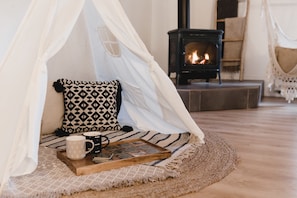 Une pièce chaleureuse avec un lit, un baldaquin blanc, un coussin à motifs, un plateau avec une tasse et un livre, et un poêle à bois en arrière-plan.