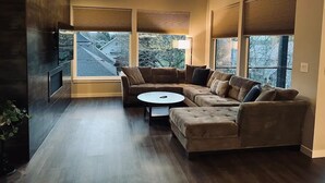 Ein modernes Wohnzimmer mit einer Eckcouch, einem runden Couchtisch und großen Fenstern mit Jalousien.