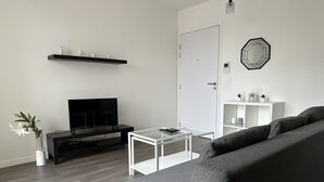 Un saló modern amb un sofà gris, una televisió de pantalla plana, una taula de centre i una taula de consol blanca amb objectes decoratius.