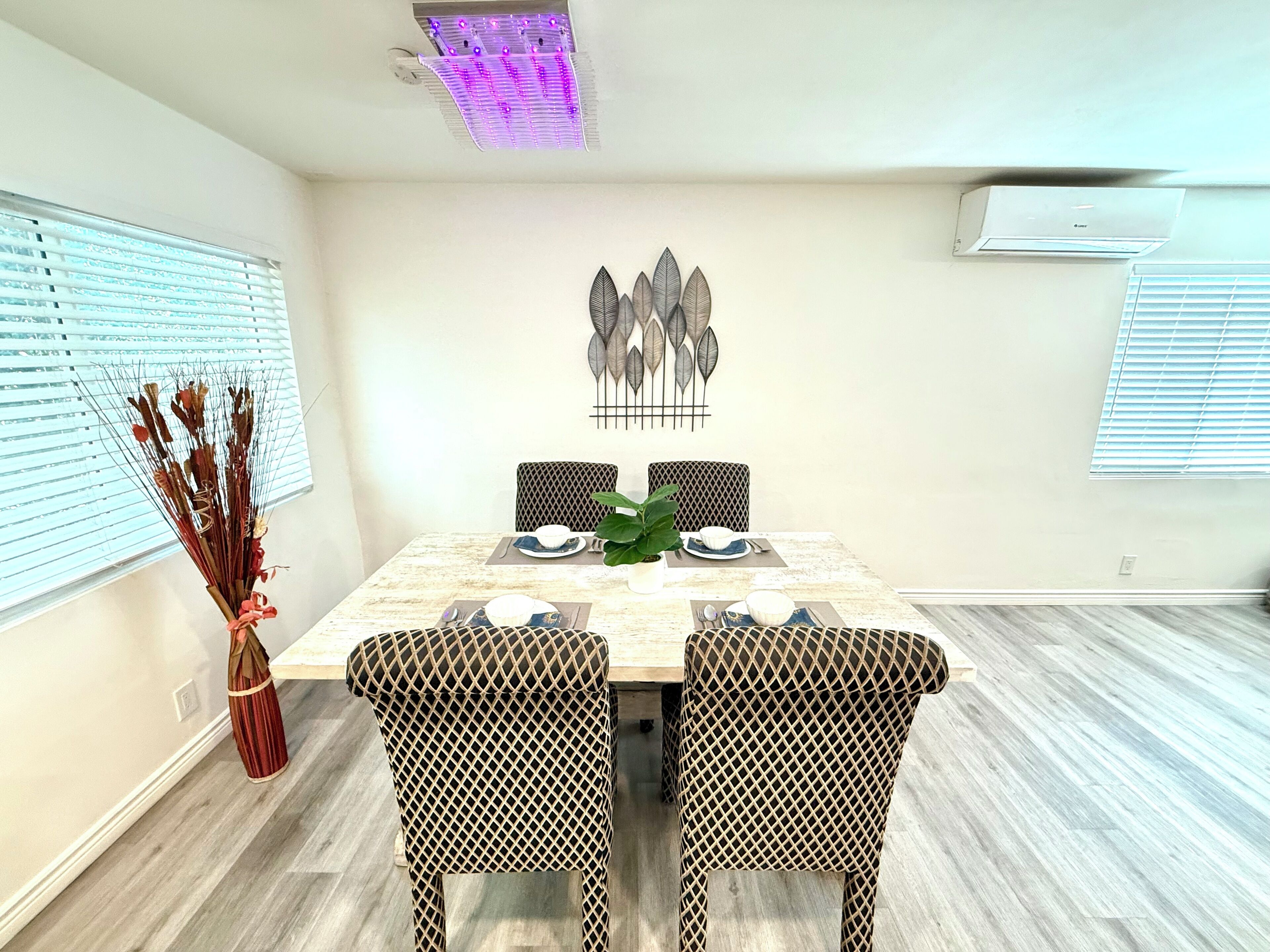 A dining area with a rectangular table, two patterned chairs, a decorative wall piece, and a vase with dried plants.