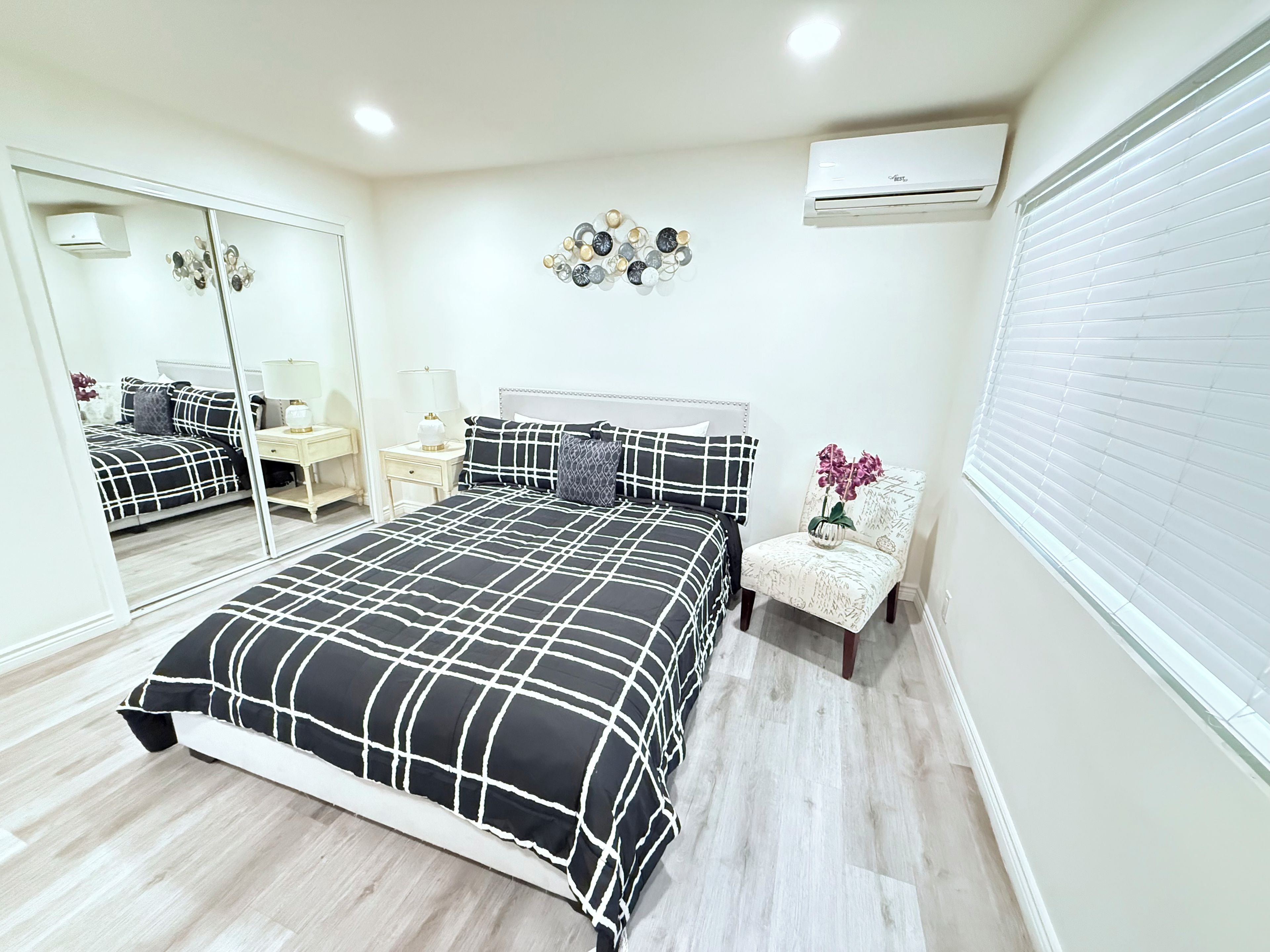 A bedroom with a bed, a chair, a mirror, a wall-mounted air conditioner, and a decorative wall piece.