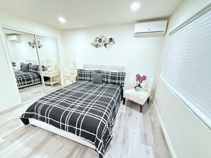 Un dormitorio con una cama, una silla, un espejo, un aire acondicionado de pared y un adorno en la pared.