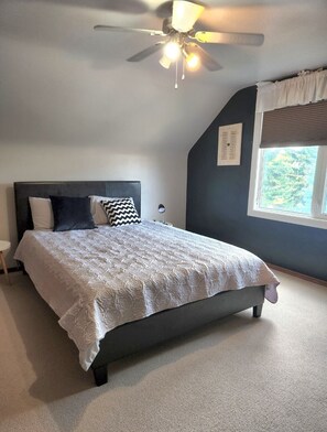 Un dormitorio con cama, ventilador de techo, ventana con persianas y mesita de noche.