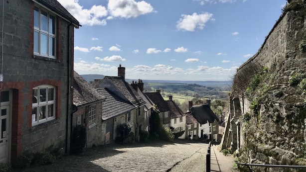 A narrow cobblestone street flanked by historic stone buildings with traditional architecture, leading to a scenic view of rolling hills. - Ramblers, Marnhull, Shaftesbury (Marnhull)