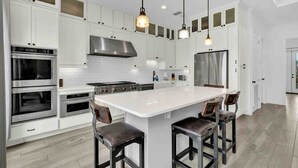 A modern kitchen with white cabinetry, stainless steel appliances, a large island with bar stools, pendant lighting, and a visible stainless steel range hood. - 5909 Havana Dr: Private Pool/Large Back Yard (South Padre Island)