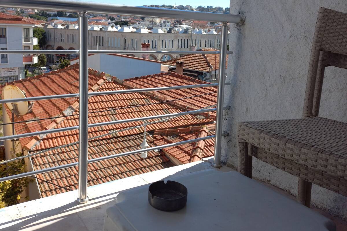 A balcony with a wicker chair and a small table, overlooking a cityscape with red-tiled roofs and white buildings.